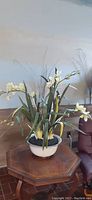 Full view of light color faux lilies with green leaves arranged in porcelain pot with gold trim. The piece sits on a wooden surface.