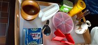 Overview of assorted kitchen items on white tray including wooden bowl, measuring spoons, ladle, decorative robin figurine, spice tins, and silicone pot holders.