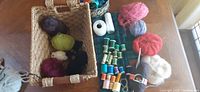 Photo showing a basket with various skeins of wool and mohair blend yarns in colors such as dark purple, green, maroon, black, white, red, pink, and grey, along with sewing thread spools and white cotton string spools on a wooden table.