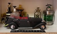 Photo showing black vintage car decanter holder with cut glass decanter and six shot glasses inside. Additional decanters including steam train and lantern style are behind.