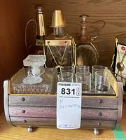 Four decorative barware decanters displayed in a wooden/sleeve barrel-style holder along with four shot glasses. The lot includes a clear cut glass decanter, a tall gold-tone Gin decanter, a pink-tinted stopper decanter, and a separate decanter inside the barrel holder.
