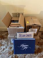 Two boxes filled with books about New York City, nautical history, and ships. Two large coffee table books in front: 'Romance of the Sea' and 'Ocean Liners Travel on the Open Sea'. Boxes labeled 'Sea Stories'.