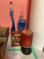 View of all four items grouped on surface against pink background. Includes two blue glass decanters, wooden book dispenser, red and gold music box.