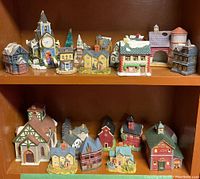 Two shelves displaying a total of 16 ceramic miniature buildings including a church, barn with silo, store, and assorted houses and cottages.