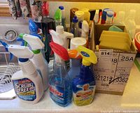 Image showing multiple spray bottles of various cleaning liquids including Scrub Free, Windex, and others. Bottles mostly white with trigger spray tops in different colors. Bottles are placed on countertop next to a sink.