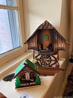 Two item lot showing a wooden cuckoo clock and nearby plastic thermometer with couple figurine, on window ledge.