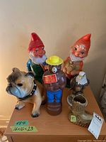 Entire lot of figures displayed on a wooden surface including plastic gnomes, Smokey Bear, ceramic bulldog, eagle, and cowboy boot container.