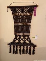 Full frontal view of black paracord macrame wall hanging with wood beads and hanging tassels, mounted on wall with wooden rods and black hanging cord. Purple ribbon tag attached to indicate ribbon-winning status.