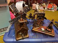 Four metal musical box sculptures arranged on a blue plastic surface; copper-toned windmill, horse and rider sculpture, and sailing ship with lighthouse among them.