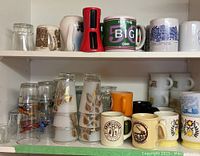 Two shelves of assorted mugs and glasses showcasing a variety of designs, including commemorative and vintage styles, alongside a red spiralizer kitchen tool