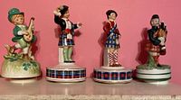 Four Scottish-themed music box figurines standing side by side on a shelf against a pink background