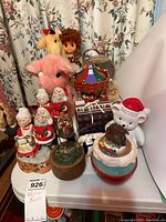 Various musical stuffed animals, holiday figurines including multiple Santa Claus playing instruments, a pink elephant plush, giraffe plush, white teddy bear with Santa hat, carousel water globe, and other Christmas decorations.