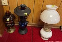 Three vintage oil lamps side by side on red carpet in front of wood paneling wall. Includes brown glass oil lamp with brass base, dark blue glass oil lamp with metal top, and white hobnail glass oil lamp with floral base.