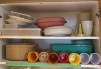 Shelves containing assorted plastic food storage containers in white, beige, and pink along with colorful drinking glasses in yellow, orange, red, and blue.