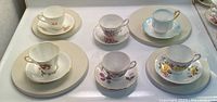 Six china tea cups and saucers displayed on circular white platforms on a white surface. Varied floral and color designs visible.