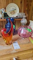 Photo showing orange Murano style vase, pink cranberry bowl, blue glass vase, and small oil lamp on wooden surface with background wall and miscellaneous items