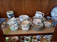 Photo showing Indian Tree pattern tea cups, saucers neatly stacked, with creamer and bowl in the background on a wooden shelf.
