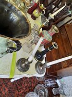Photo showing assorted candle holders on a white table, including silver-toned holders with white candles, black candle holder, and decorative brass-like vase with printed design.