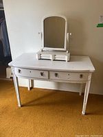 Front and side angle view of the white painted wood desk showing the three drawers and curved front edge, surface has scratches and wear marks.