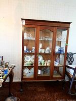 Frontal view of the wood display cabinet showing three glass doors and internal shelves with items. Dark polished wood and decorative molding on top visible.