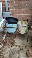 Photo of two plastic planters on a rusted metal stand against a brick wall, one light blue with black rim, the other cream-colored with matching saucer.