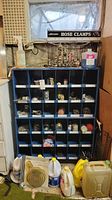 Full front view of blue metal cabinet with 30 cubbies holding assorted plumbing and hardware parts, with containers of fluids on the floor in front.