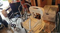 Black wheelchair and white shower chair with adjustable legs in a garage setting with boxes and a bicycle in background.