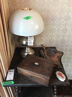 Converted oil lamp with white cracked plastic shade, copper crumb catcher, wooden box, and small white ceramic dish on a carved wooden table.