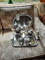 Full coffee service set displayed on a floral patterned chair featuring all coffee pots, creamer, sugar bowl on the smaller tray