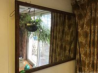 Rectangular wall mirror with dark wood frame hung on a wall. Reflection shows curtains, a door, and plants.