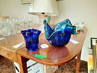 Two blue Murano glass bowls displayed on a wooden table along with glassware and lamp in background.