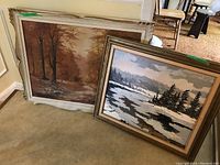 Two framed oil paintings on canvas leaning against a wall indoors on carpet. One painting shows a woodland scene with tall trees and an earth-tone color palette in an ornate white frame. The other depicts a winter river or lake with dark trees and snow in a wood-toned frame.