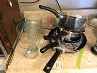 Overview of stacked T-Fal pots and pans wrapped in plastic, glass pitcher and glasses beside them on beige countertop.