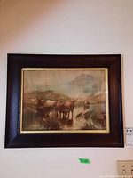 Photo of the framed print showing cattle standing in water with a mountain landscape background, framed in dark brown with gold border.