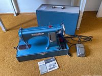 Full view of Kenmore electric sewing machine, foot pedal, carrying case behind, and manual on floor