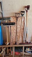 Photo showing three snow shovels with wooden handles and plastic blades leaning against a wall, wooden stool and tile table top behind