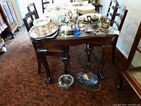 Photo showing dining table with multiple silver plated and glass serving ware items including trays, serving pot with lid, creamer, teapot, wine cup, beer stein, salt and pepper shakers, and stacked plates.