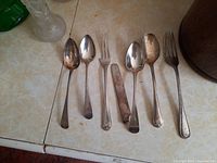 Full set of 8 sterling silver flatware pieces laid out including spoons, forks, and a butter knife with varied handle styles and tarnishing.