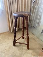Full view of wooden stool showing round seat and turned legs on carpeted floor.