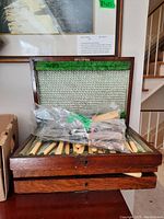 Wooden case open showing cutlery arranged in rows with green felt interior and some items bagged for protection