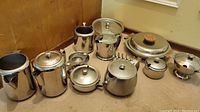 Wide view showing nine stainless steel vintage metalware pieces including pots, sugar bowls, and covered vegetable bowl with wood handles.