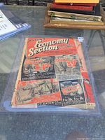 Photo showing the full front cover of the 1936 Eaton's Economy Section catalog laid flat within a clear hard plastic holder on a reflective surface with other items in the background.