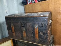 Front view of the antique metal and wood humpback trunk showing worn black paint, rusted metal latches and hardware, and wooden vertical slats on the front.