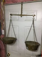Full view of vintage solid brass balance scale with two chains and pans hanging from a metal bar mounted on door.