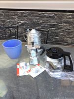 Bialetti coffee maker set on outdoor table includes aluminum stovetop coffee maker, glass pot, blue planter, and aluminum ice bucket.