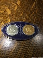 Top-down view of two Ontario Centennial Confederation Commemorative Coins, silver and gold colored, housed in a clear oval plastic case with blue fabric backing on a wooden table.