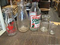 Five clear glass dairy bottles on a wooden table, including 3 milk bottles with different logos and 2 small creamer bottles.