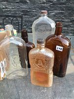 Group of five old liquor and wine bottles outdoors on a table, showing a variety of shapes and colors including clear and amber glass.