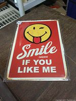 Photo showing full metal sign with red background and yellow winking smiley with tongue out graphic, text 'Smile if you like me'. The sign is in plastic with edges showing rust-like details.