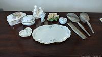 Full lot overview showing all porcelain trinket boxes, figurines, Lenox tray, and silver-tone brush set.